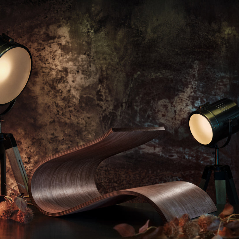 A sculptural wooden table lamp with a curved, wave-like wood grain finish, styled on a dark surface with two studio spotlights against a textured dark background.
