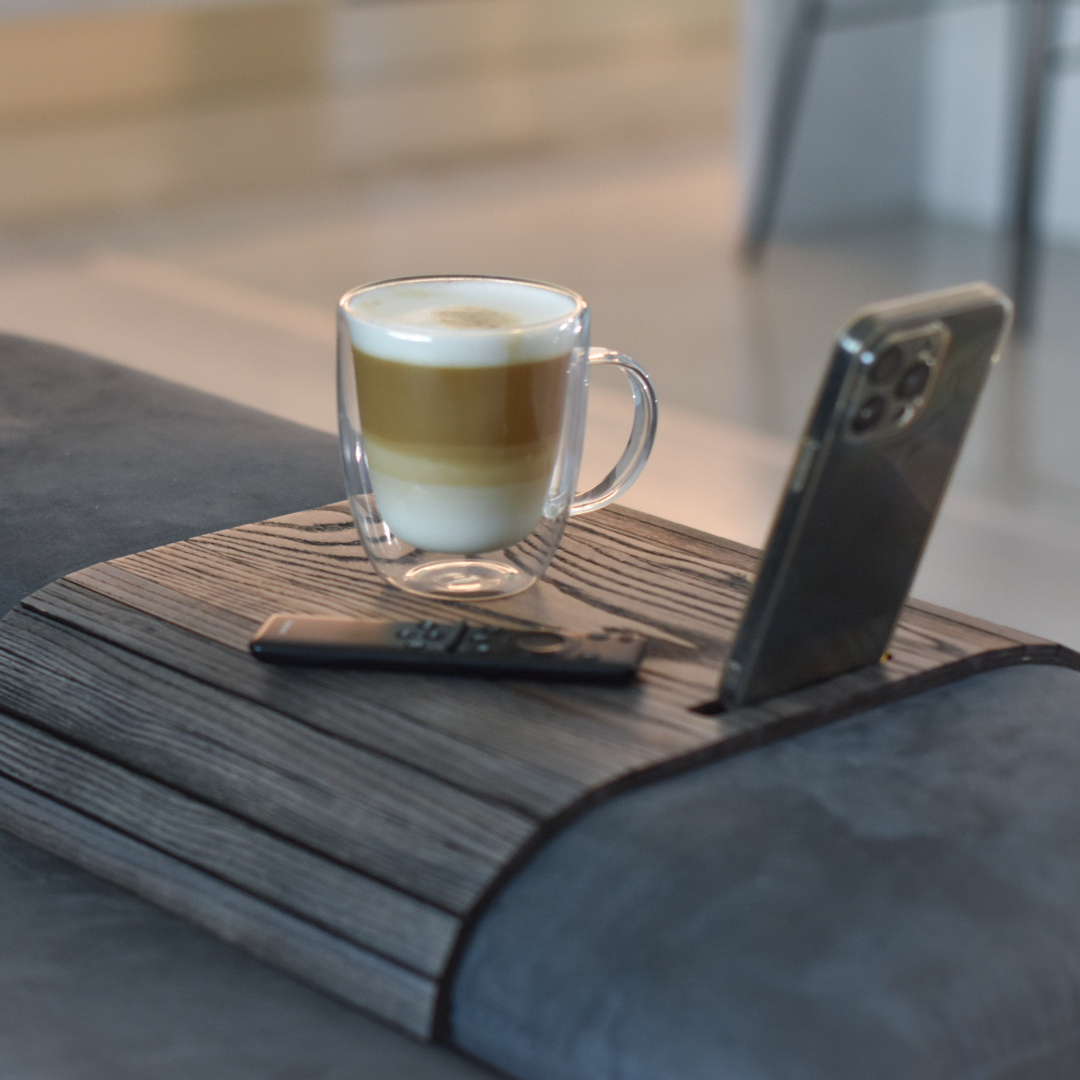 brown wooden sofa arm tray table on sofa arm with tv remote and coffee cup on it