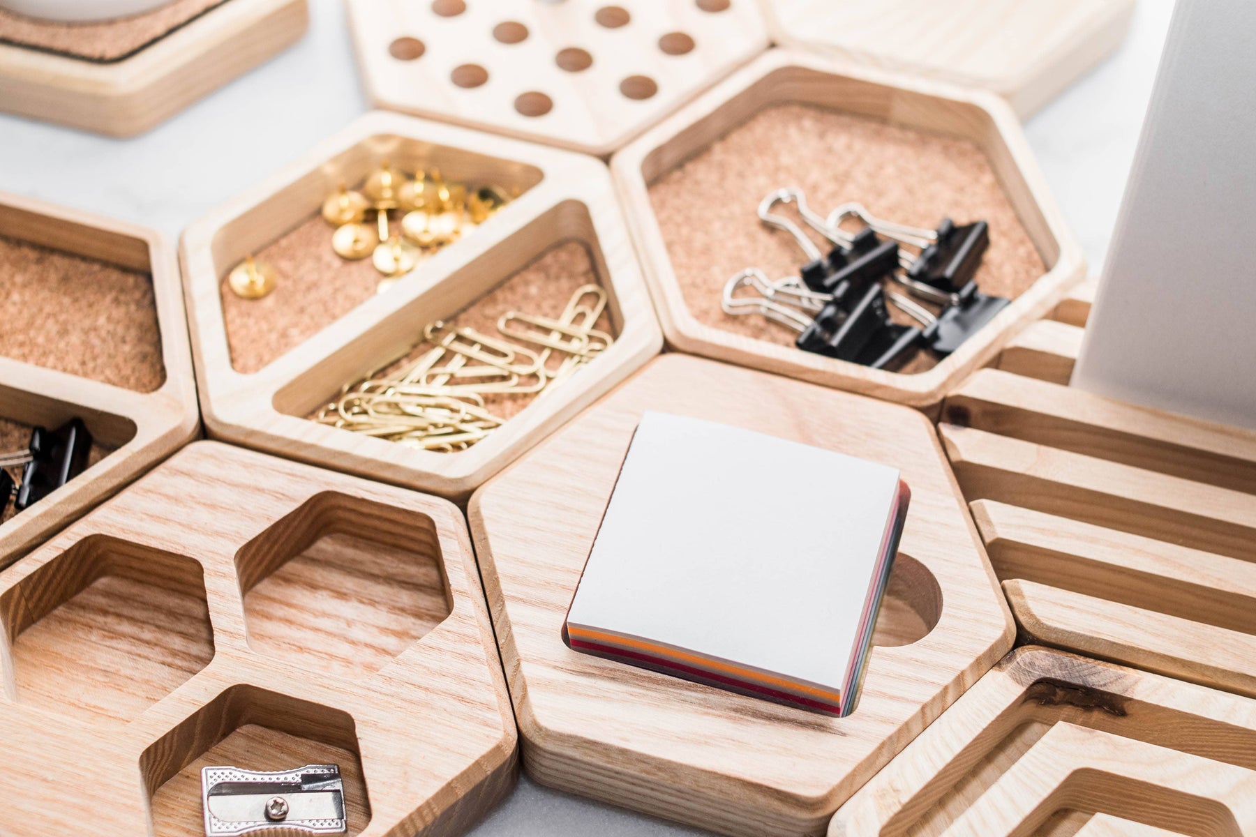 Hexagonal cork inlay organizers with wooden compartments, holding office supplies such as paper clips, thumbtacks, binder clips, and sticky notes. A modular and stylish desk accessory for organized workspaces.