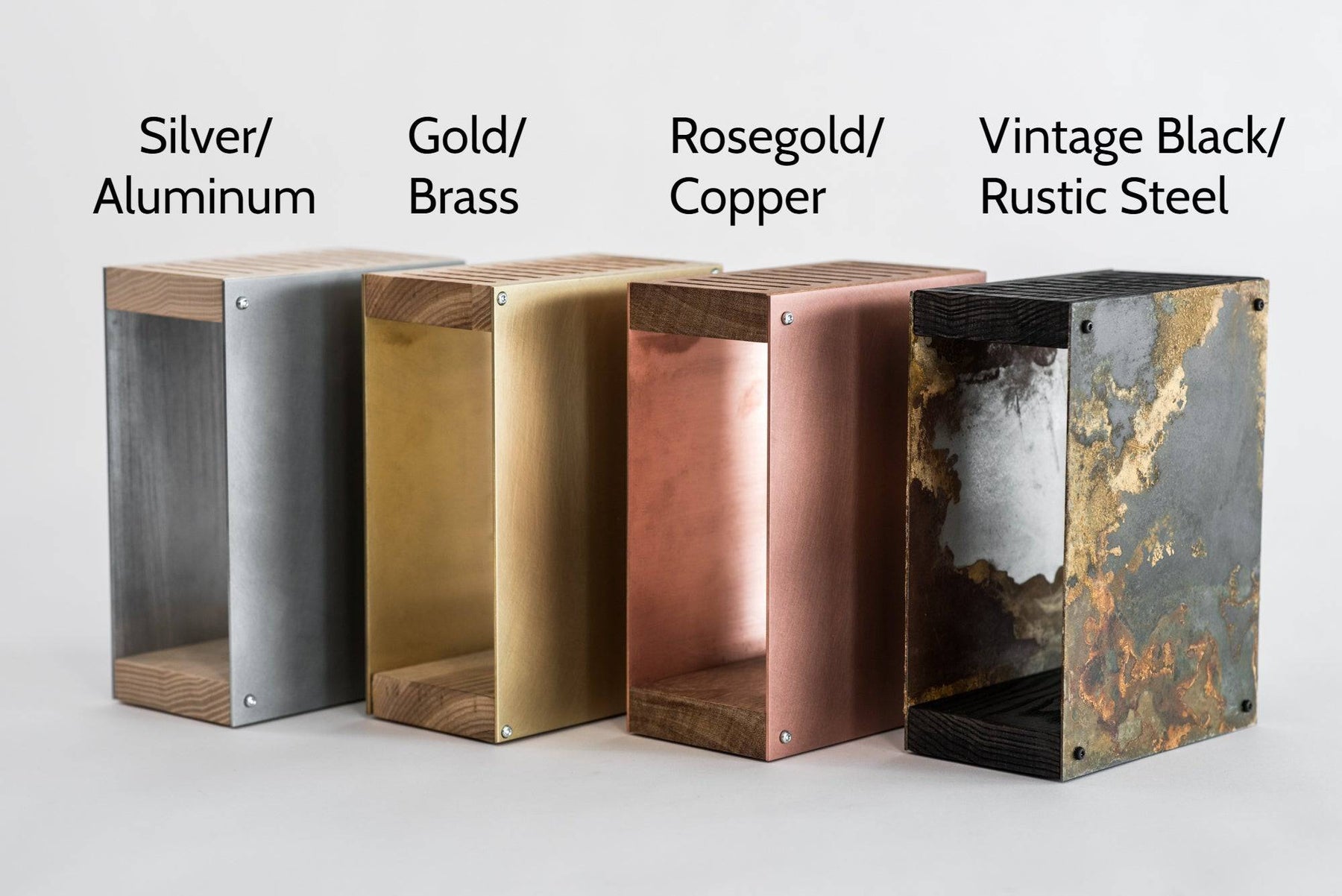 Four metal knife blocks in different finishes: silver/aluminum, gold/brass, rosegold/copper, and vintage black/rustic steel. Each block has a wooden top and base, displayed side by side against a plain background.