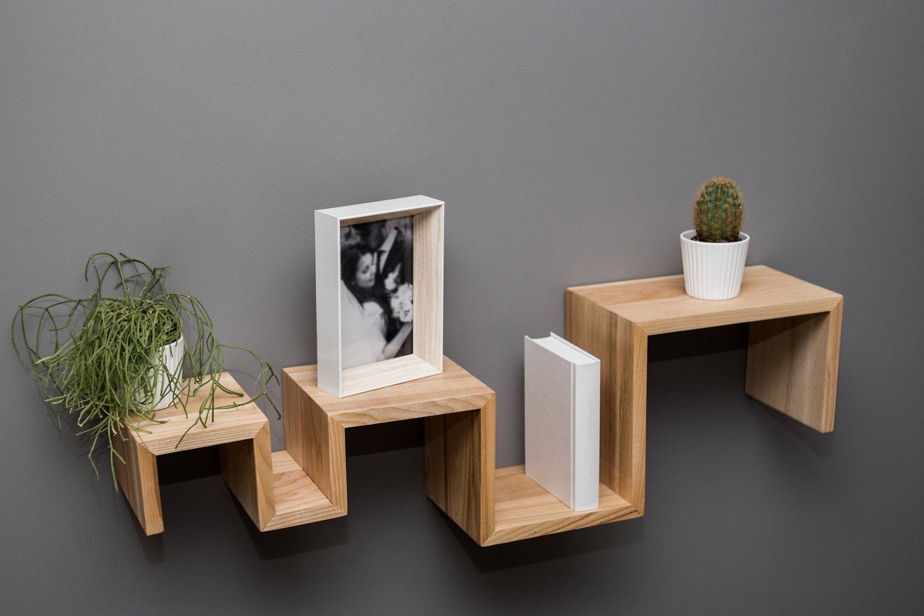 Three-step wooden shelf with a natural wood grain finish, mounted on a gray wall. The shelf holds a potted plant, a framed photo, and a book