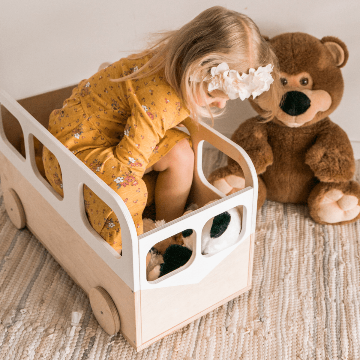 A young girl sits inside a natural wood toy bus with white accents and round wooden wheels, placed on a woven rug with a stuffed bear nearby.