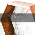 Hexagonal shelf made of rust-colored metal and natural wood, featuring a glass vase with dried plants. The Ewart Woods logo and the slogan "Make Simple Beautiful" are overlaid on a semi-transparent black strip.