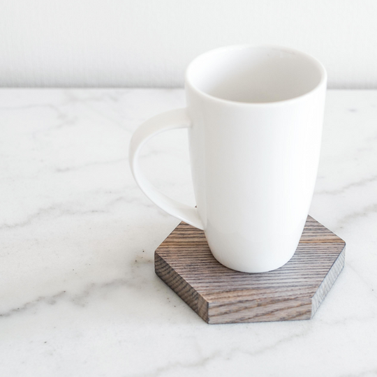 A white ceramic mug resting on a dark wood-finish hexagonal coaster, set against a light marble surface, creating a clean and minimalist aesthetic.