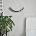 Front view of a dark brown curved toilet paper holder with a hidden mounting system on a tile bathroom wall, near a modern toilet. A plant adds greenery to the minimalist bathroom design.