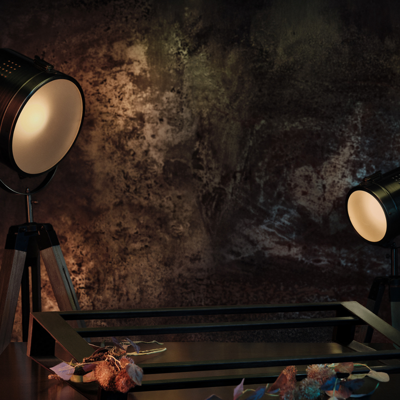Industrial-style black floor lamps with warm white light on a dark rustic textured wall, with a black metal footwear holder in the foreground and dried flowers at the bottom.
