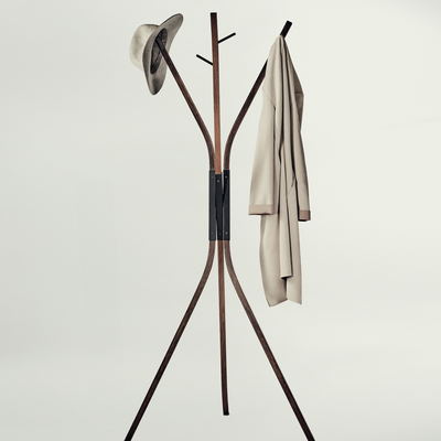 A modern wooden coat rack with three legs and curved top arms, shown against a plain light gray background. A hat is hanging on the left hook and a light beige coat is hanging on the right.