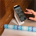 A smartphone placed on a white cell phone holder, positioned on a wooden surface next to a roll of patterned wrapping paper, with a hand interacting with the phone&