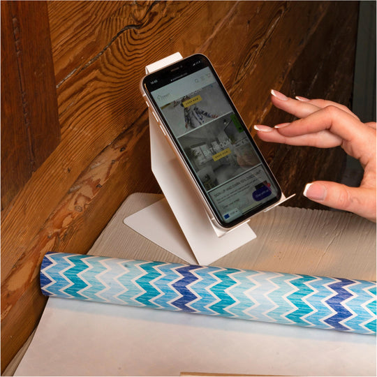 A smartphone placed on a white cell phone holder, positioned on a wooden surface next to a roll of patterned wrapping paper, with a hand interacting with the phone&