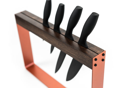 Angled view of a modern knife holder with a dark wooden top, vibrant orange metal frame, and visible black screws, holding four black-handled knives in a single vertical slot.