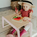 Young child in a sunhat and coral dress plays with wooden toy food at a pastel pink and natural wood activity table, wearing bright pink rain boots.