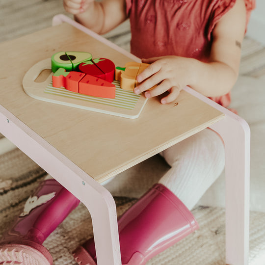 Child in a red dress and bright pink boots interacts with wooden play food on a natural wood and blush pink activity desk.