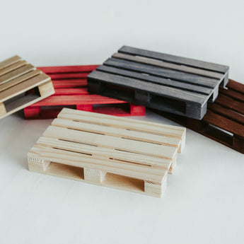 A variety of handmade wooden pallet coasters in different colors, including natural wood, red, gray, and dark brown, arranged on a white surface.