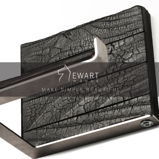 Close-up of a burned ash wood and stainless steel toilet paper holder with the EWART WOODS logo and the slogan MAKE SIMPLE BEAUTIFUL overlaid. The sleek metal bar and textured wood combine for a stylish, functional design.