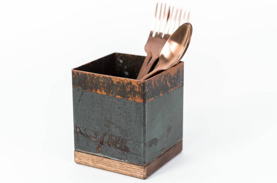 Square utensil holder with a rustic copper finish and wooden base, containing copper-colored forks and a spoon. The holder is placed against a plain white background.