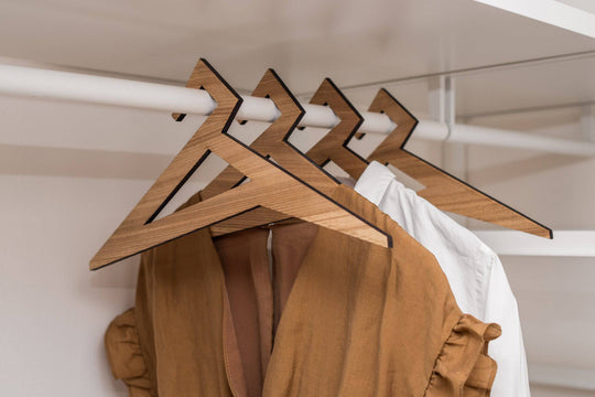 Wooden hangers in paldau finish with a geometric design, holding a brown dress and a white shirt in a closet.