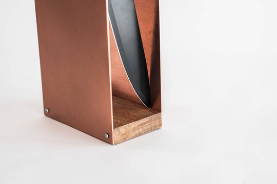 Close-up of a rose gold knife block with a smooth metal finish and wooden base, featuring a side slot with a partially visible knife blade. The block is displayed against a plain white background.