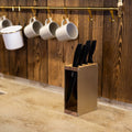 Gold-colored knife block with a wooden top and base, holding black-handled knives. The knife block is placed on a kitchen countertop with a rustic wooden wall in the background.