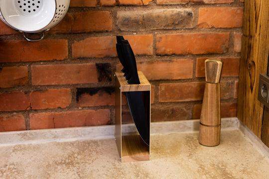 Wood and metal knife block with black-handled knives on a kitchen countertop, set against a rustic red brick wall. A wooden pepper grinder and a metal colander hanging from the wall are visible in the background.