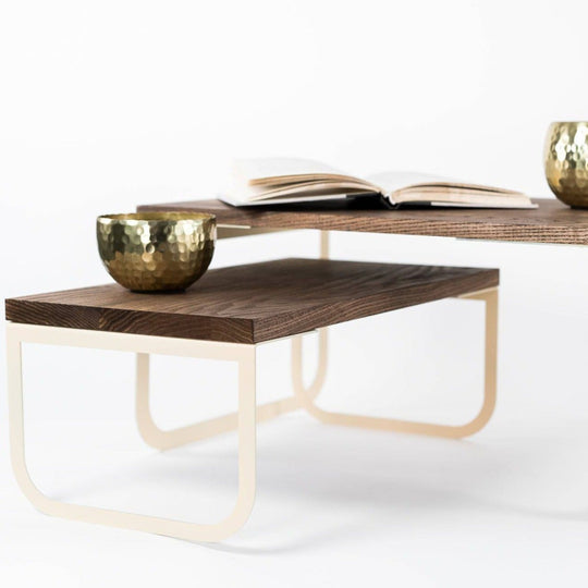 Modern serving tray with dark wood surfaces and cream-colored metal legs, featuring two gold cups and an open book, displayed on a white background.