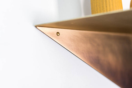 Close-up of the corner of a triangular metal wall shelf with a visible screw, showcasing the brushed copper finish.