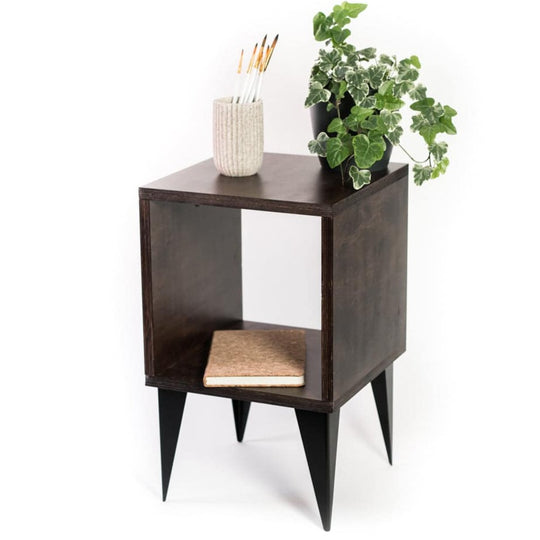 Dark-stained wooden side table with a square open compartment, black tapered legs, and a minimalist design. Decorated with a plant, a ceramic container holding paintbrushes, and a cork notebook placed inside