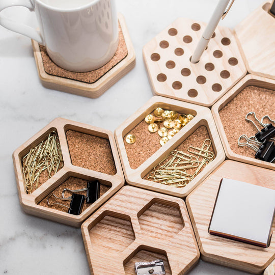 Hexagonal wooden desk organizers with cork inlays, storing office supplies such as paper clips, thumbtacks, and binder clips. A matching wooden coaster with a mug and a pen holder add functionality and style to the setup.