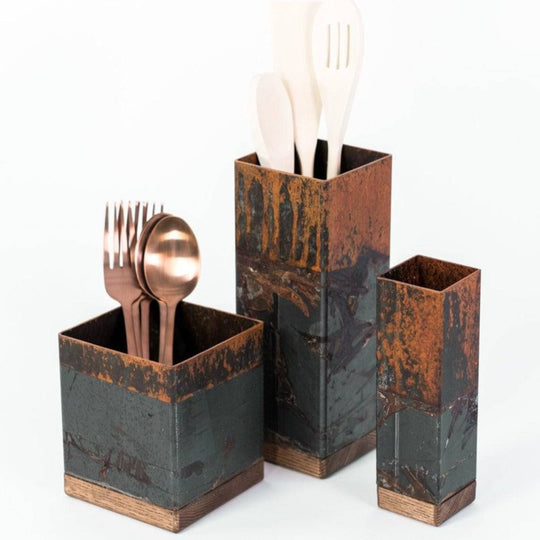 Three rustic, metal utensil holders of varying heights with a weathered copper finish. One holder contains copper-colored forks and spoons, another holds wooden utensils, and the smallest holder is empty.