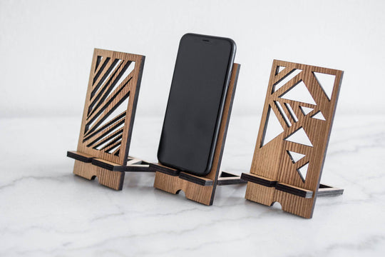 Wooden phone stand set in walnut finish with a geometric pattern, holding a phone in the center, placed on a smooth surface. The stands are designed with modern, angular shapes on each side.