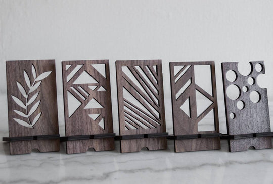 Wooden phone stands in walnut finish with unique geometric cutout designs:  Leaf pattern, Abstract triangles, Diagonal lines, Geometric shapes, Circular cutouts. Each stand holds a phone at an angle on a smooth surface.