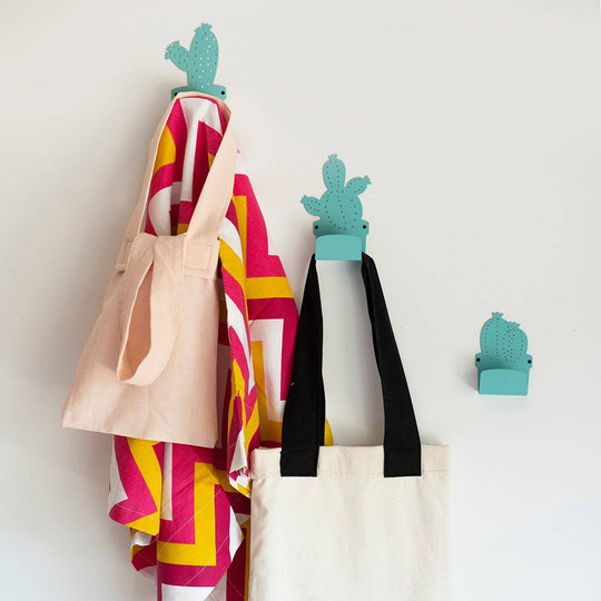 Three teal metal cactus-shaped wall hooks holding a pink tote bag, a colorful towel, and a black and white tote bag against a white wall.
