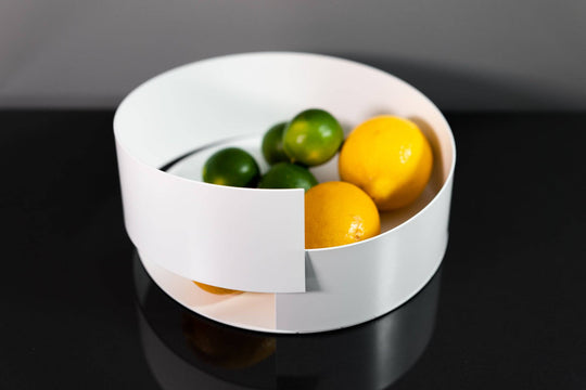 Modern white metal bowl with a sleek, overlapping design, holding vibrant lemons and limes, displayed on a reflective black surface.