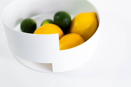 Modern white metal bowl featuring a clean, asymmetrical design, filled with bright citrus fruits, displayed on a minimal white background.