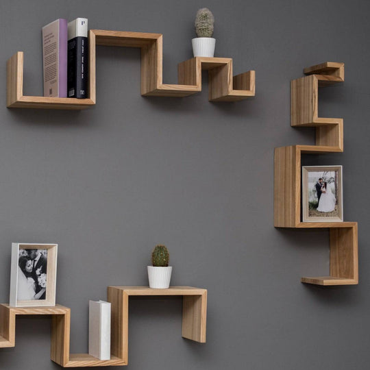 Three floating wooden shelves with geometric, zigzag designs mounted on a dark gray wall. The shelves hold a variety of items, including books, small potted cacti, and framed photographs.