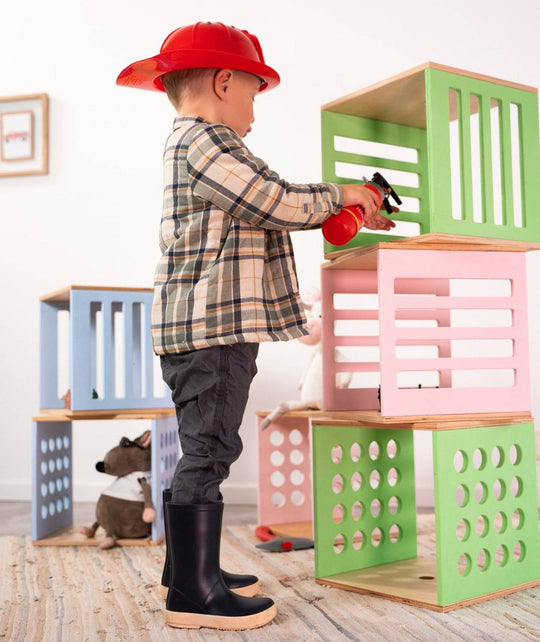 A child wearing a firefighter helmet and boots holds a toy fire extinguisher aimed at a stack of colorful wooden blocks with various cut-out designs, including slats and circular holes.