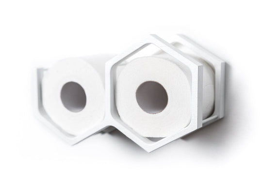 Close-up of a wall-mounted white mini honeycomb toilet paper holder, displaying two rolls of toilet paper. The holder features metal bars for support and a geometric design with textured wood.