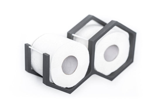 Close-up of a wall-mounted gray mini honeycomb toilet paper holder, displaying two rolls of toilet paper. The holder features metal bars for support and a geometric design with textured wood.